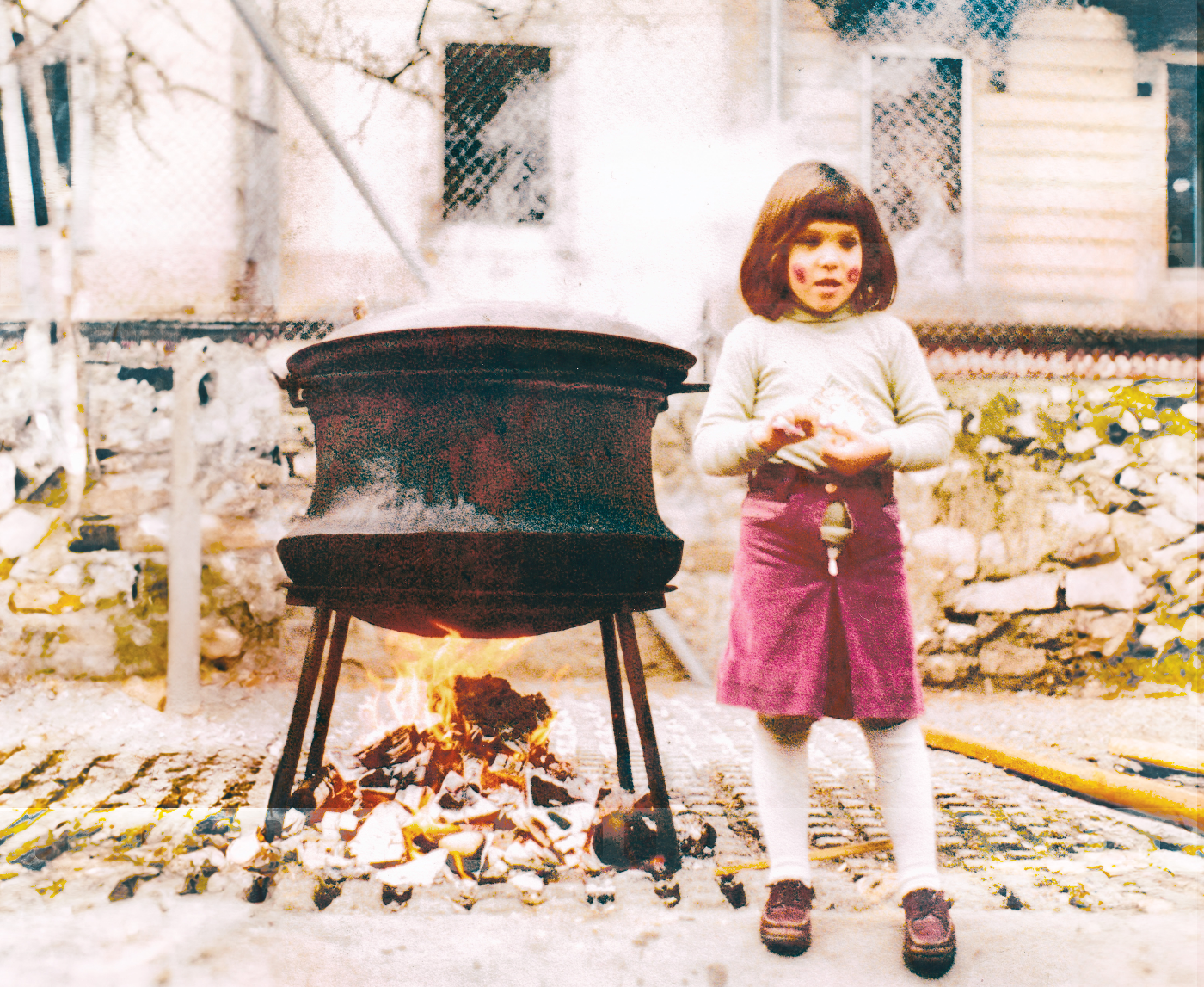 Une enfant près d’une marmite sur un feu de bois, devant un mur de pierre.